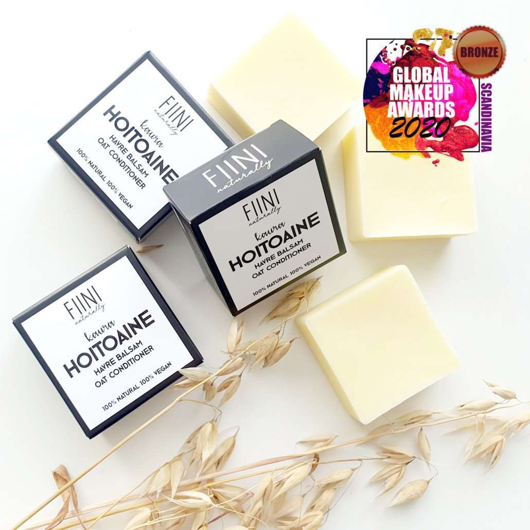 Fiini Naturally Kaura Hoitoaine; neljä hoitoainepalaa pakkauksineen ja kuivattua heinää aseteltuna valkoiselle taustalle, oikeassa yläkulmassa Global Makeup Awards 2020 Bronze -mitalimerkki; suorakulmaiset pakkaukset selkeällä "Fiini"-brändäyksellä yksinkertaisella groteskikirjasimella; palat pyöreitä ja sileän mattapintaisia beigen ja vaaleankeltaisen sävyissä; kuvattu ylhäältä, ei ihmisiä kuvassa.