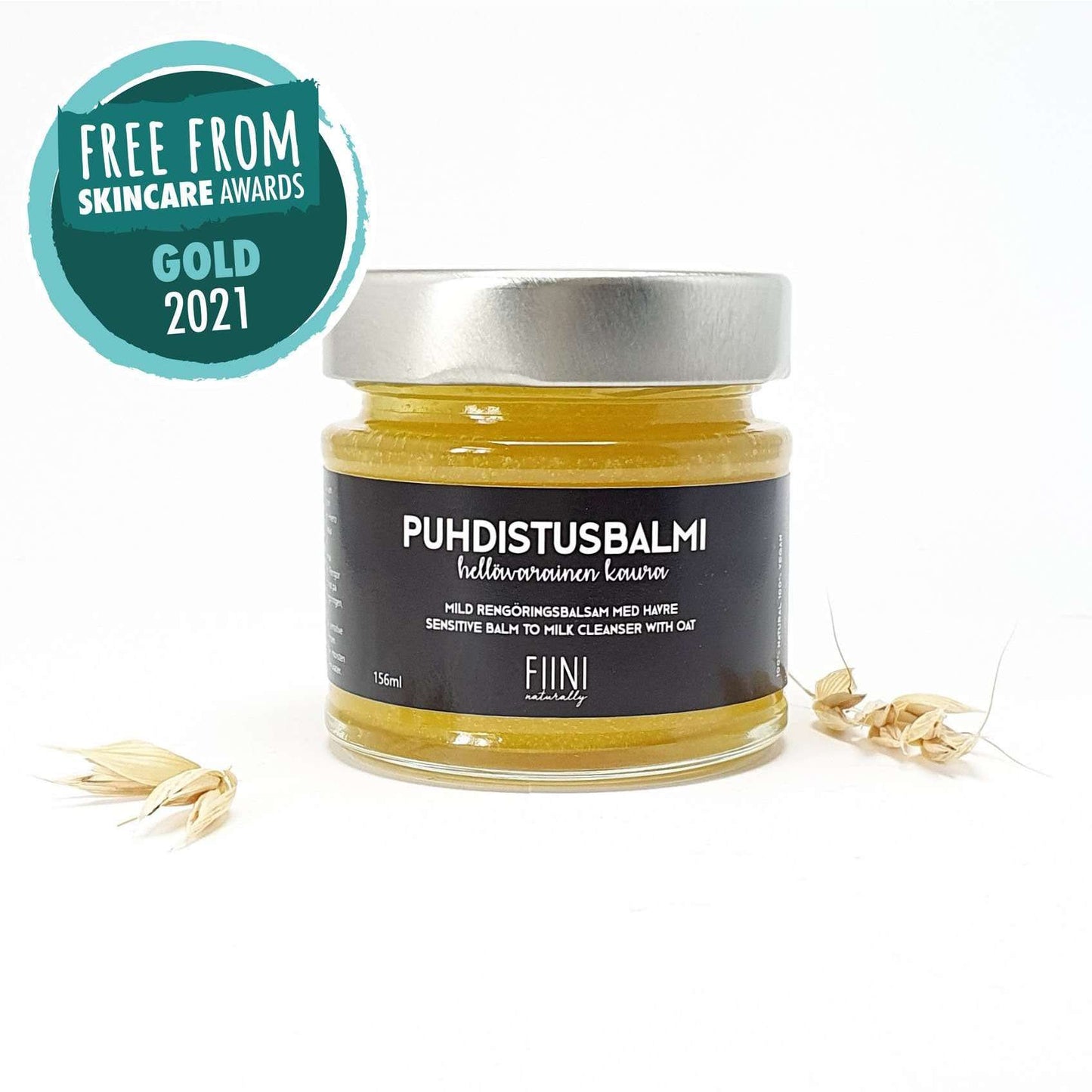 Fiini Naturally Puhdistusbalmi Hellävarainen Kaura; lasipurkki kaurajyvien vieressä, korostaen kauran luonnollisia hyötyjä; vasemmassa yläkulmassa Free From Skincare Awards Gold 2021 -mitalimerkki; purkki sylinterinmuotoinen, valkoinen etiketti mustalla groteskitekstillä, kansi metallinen ja mattapintainen; valkoinen tausta, ei ihmisiä tai käsiä, tasainen studiovalaistus.