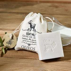Olivos Vuohenmaitosaippua; suorakulmainen vaaleanbeige saippuapala kohotetulla vuohikuviolla, vieressä luonnonvärinen narusuljettu puuvillapussi tekstillä "Goat Milk Olive Oil Soap"; lisäksi pieni valkoinen astia ja neliönmuotoinen etiketti kuvitetulla vuohella; kaikki aseteltu vaakasuorille puulankuille; ei ihmisiä tai käsiä kuvassa.
