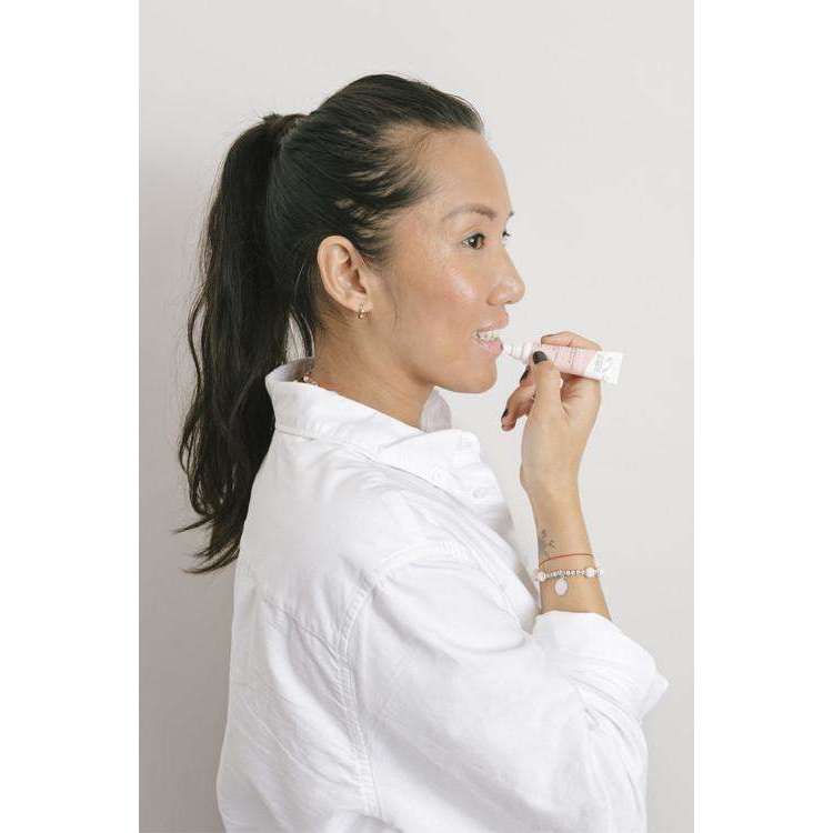 Alga Cicosa Reparative Lip Balm Korjaava Huulivoide; poninhäntäinen nainen valkoisessa paidassa levittää huulivoidetta pienestä lieriömäisestä tuubista; tuubi sileäpintainen, todennäköisesti muovia ja vaaleansävyinen, etikettitiedot eivät ole luettavissa; nainen pitelee korkitonta tuubia huultensa lähellä oikealla kädellään; tausta vaalea ja yksinkertainen; kuva on lähikuva tai puolivartalokuva ilman muita esineitä.