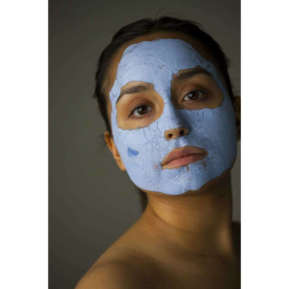 BRUNS Products Nº101 Blue Clay Savinaamio; henkilö pitkin tummin hiuksin ja paljain hartioin katsoo kameraan, kasvoilla kuivunut ja halkeillut sininen savinaamio; naamio mattapintainen ja epätasainen halkeamien vuoksi; rajattu tiiviisti kasvoihin ja yläosaan; tausta neutraali.