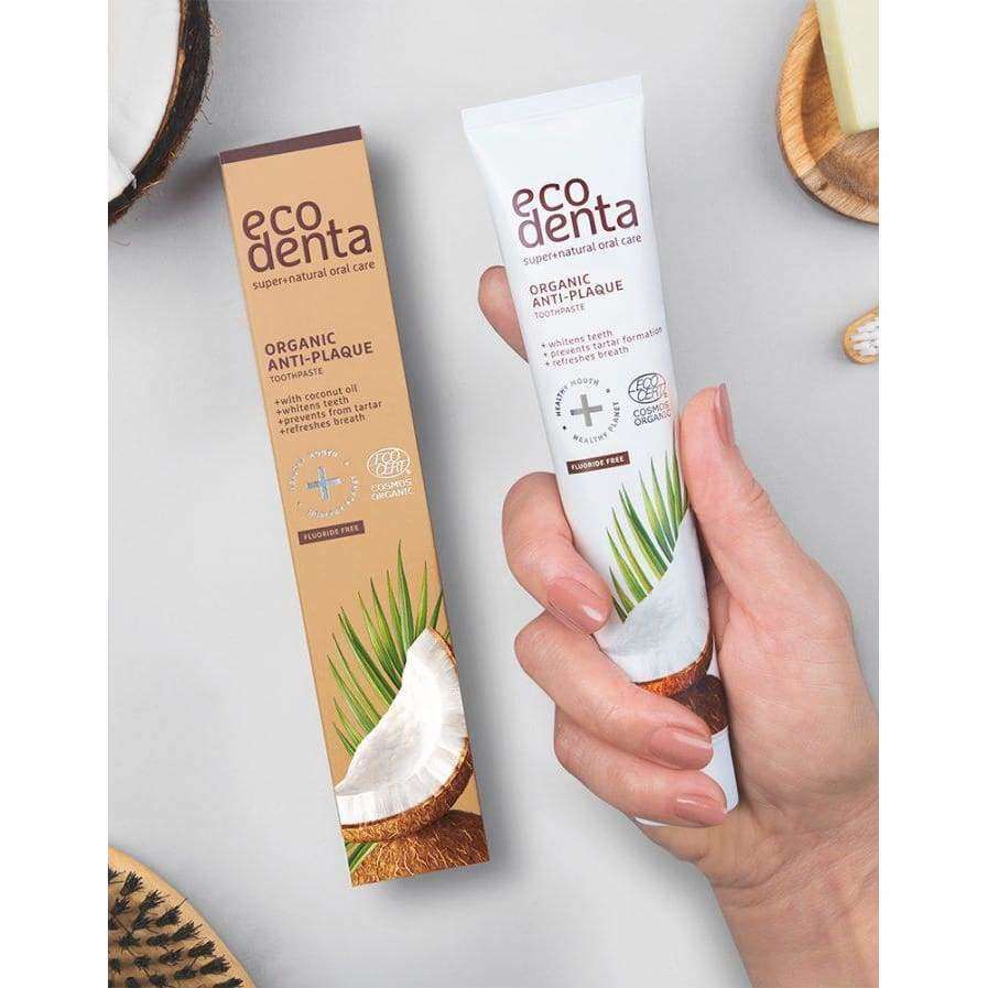 Ecodenta Anti-Plaque Hammaskiveä Ehkäisevä Hammastahna; käsi pitelee Ecodenta Organic Anti-Plaque -hammastahnatuubia vieressä olevan rasian kanssa, molemmissa kookos- ja kasvikuvitusta harmaalla pinnalla ekologisten puisten kylpyhuonetarvikkeiden ympäröimänä; tuubi valkoinen mattapintainen, vihreä ja ruskea grafiikka ja groteskifontti tuotenimelle; pakkausrasia vastaa tuubia väriteemaltaan ja typografialtaan.