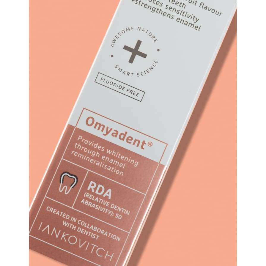 A close-up of Om yadent toothpaste packaging, highlighting it as fluoride-free and designed for whitening and enamel strengthening, with RDA 50 and created in collaboration with Iankovitch. This vegan formula showcases a commitment to both oral health and conscious ingredients. Rectangular cardboard box and tube displayed; both have a white base color with green accents, matte finish, modern sans-serif typography, flat-lay on a plain background; no people or hands present. Brands and claims are clearly