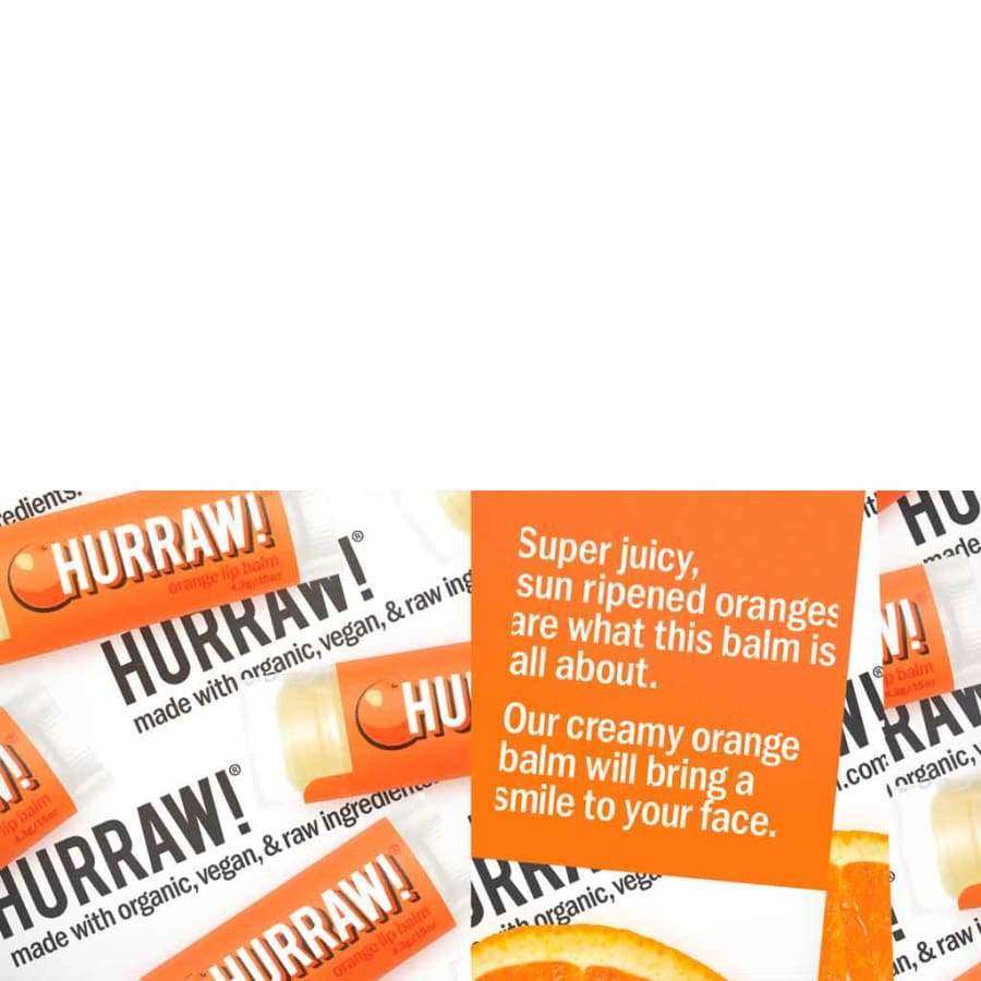 Hurraw! Huulivoide Appelsiini -tuubeja aseteltuna oranssin kyltin ympärille, oranssit mattaetiketit ja valkoiset korkit; lihavoitu groteskiteksti, kyltissä teksti aurinkokypsistä appelsiineista