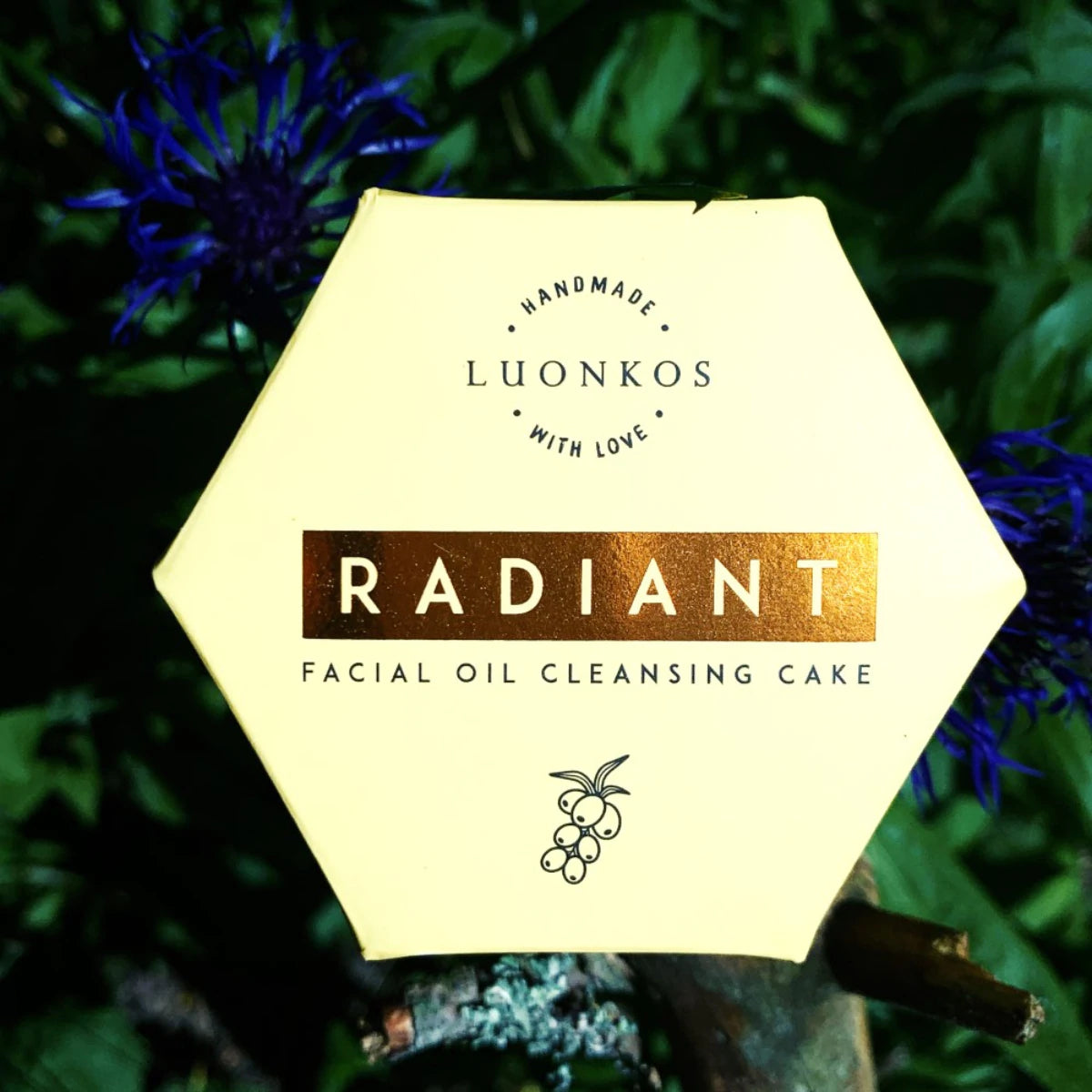 Luonkos Radiant Facial Oil Cleansing Cake -laatikko, keltainen kultaisin korostuksin, pidettynä ulkona vihreän kasvillisuuden ja violetin kukan ympäröimänä; pahvipakkaus mattapinnalla; lihavoitua tekstausvapaata etikettitekstiä; ei ihmisiä näkyvissä, laatikkoa pitää näkymätön käsi; laatikon kulmat terävät; ympäristöystävällinen pakkaus korostaa luonnollista kauneutta.