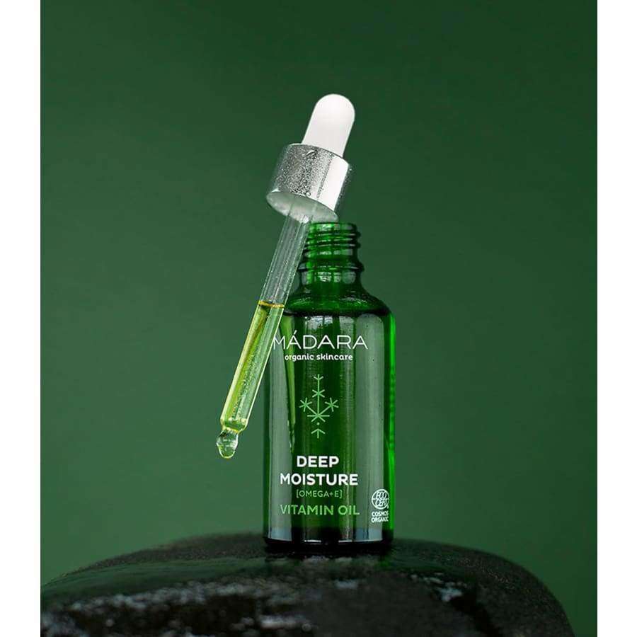 Madara Deep Moisture Vitamiiniöljy; vihreä lasipullo tekstillä "Madara Deep Moisture Omega-3-6-7-9 Vitamin Oil", tiputin pullon päällä, sisältää ravitsevaa keltaista öljyä, kuvattu tummanvihreää taustaa vasten; pullo lieriömäinen kiiltäväpinnalla; tiputin kirkas hopeanvärisellä metallikorkilla; etuetiketin typografia valkoista ja yksinkertaisen groteskia; ei ihmisiä tai käsiä kuvassa.