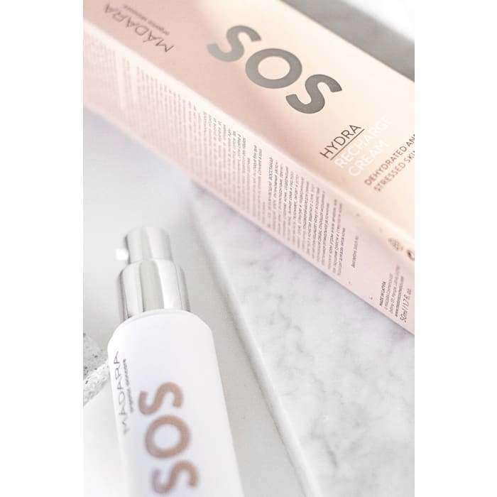 MÁDARA SOS Hydra Cream kasvovoide -pullo, valkoinen sylinterinmuotoinen, mattapinta, lihavoitu groteskiteksti "SOS HYDRA", marmoripinnalla; vieressä vaaleanpunainen suorakulmainen pakkaus samalla bränditeksillä