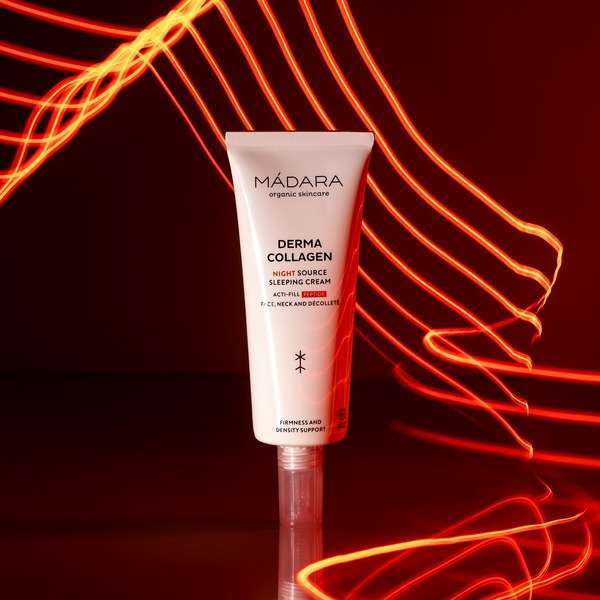 Madara Derma Collagen Night Source Yövoide; tuubi Mádara Derma Collagen Night Source Sleeping Cream -tuotetta pystyssä tummanpunaisella alustalla, ympärillä kollageenia korostavia punaisia valojuovia yhteensopivaa punaista taustaa vasten; tuubi metallinhohtoisen pinkki kiiltäväpinnalla, valkoinen groteskiteksti näyttää tuotenimen; ei ihmisiä tai käsiä kuvassa, tasainen valaistus ja keskitetty sommittelu.