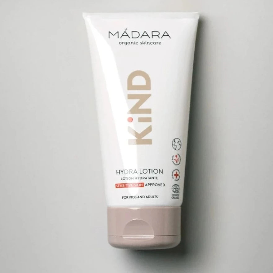 Madara KIND Hydra Lotion vartaloemulsio; valkoinen tuubi beigein korostuksin, mattapintainen ja lieriön muotoinen läppäkorkilla, tarkoitettu herkälle iholle lapsille ja aikuisille; yksinkertainen musta groteskiteksti englanniksi; pystyssä tasaisen vaaleanharmaalla taustalla.
