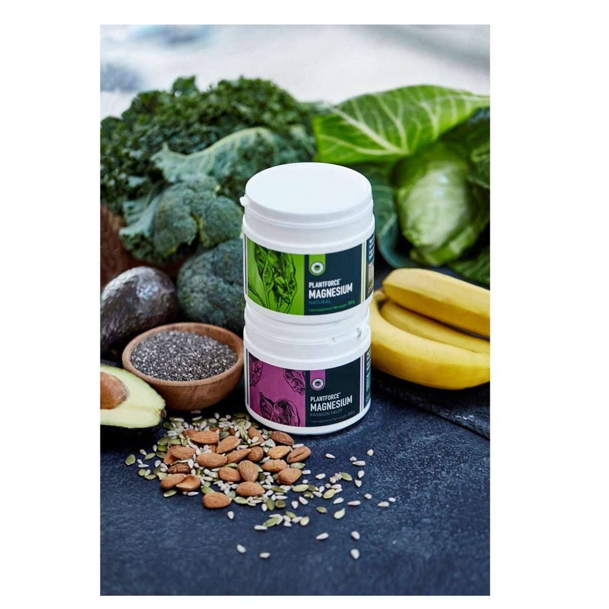 Plantforce Magnesium Maustamaton; kaksi purkkia tekstillä "Magnesium" pinottuna sileälle alustalle, mattapintaisina yksinkertaisella groteskikirjasimella; ympärillä lehtivihanneksia, banaaneja, parsakaalia, avokadopuolikkaita, chiansiemeniä ja erilaisia pähkinöitä; kirkkaasti valaistu pöytäasetelma, ei ihmisiä tai käsiä kuvassa.