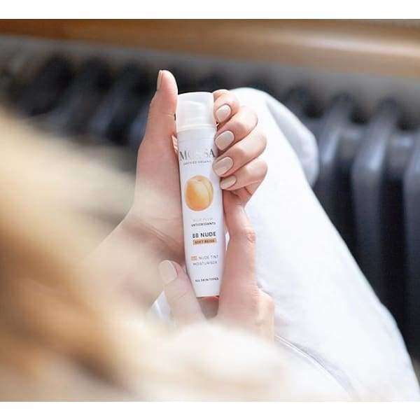 Henkilö pitelee Mossa Skin Perfector Sävyttävä BB-voide -pulloa molemmin käsin, valkoinen sylinterinmuotoinen mattapullo persikkakuvituksella; tuotenimi luettavissa groteskitekstillä, tausta pehmeästi sumennettu