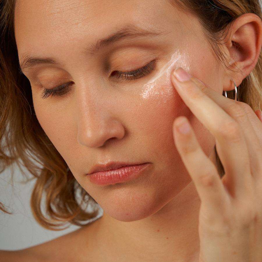 Novexpert VITAMIN C Radiance Lifting Eye Contour; lähikuva henkilöstä levittämässä silmänympärysvoidetta oikean silmän alle sormella; hiukset olkapään mittaiset, korvassa hopeinen renkaankorvakoru; voide annosteltu pienestä mattapintaisesta tuubista, väriltään valkoinen tai luonnonvalkoinen; ei näkyvää etikettitekstiä; tausta neutraali.