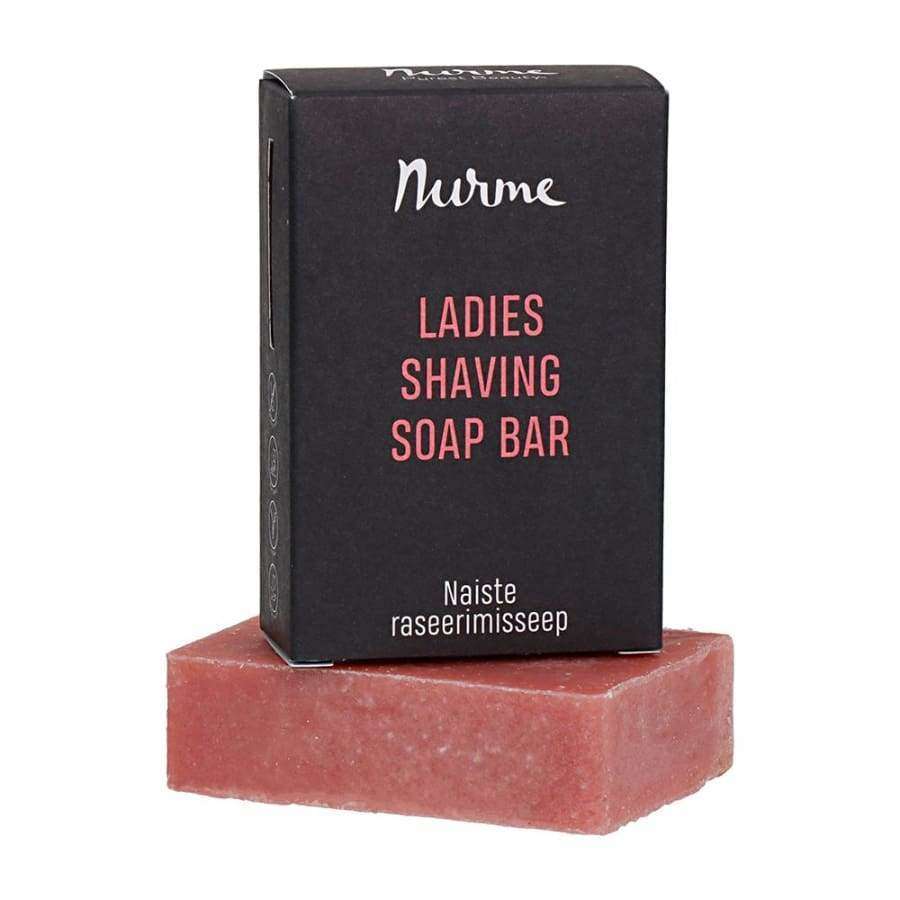 Nurme Ladies Shaving Soap Bar -parranajosaippua; suorakulmainen vaaleanpunainen saippuapala, jonka päällä musta rasia; rasiassa tekstiä myös toisella kielellä, korostaen käyttöä parranajosaippuana; saippua mattapintainen sileine reunoineen; rasian typografia valkoinen ja lihavoitu brändi- ja tuotenimelle, pienempi fontti lisätiedoille; vaalea tausta, ei ihmisiä tai rekvisiittaa; kuvattu ylhäältäpäin.