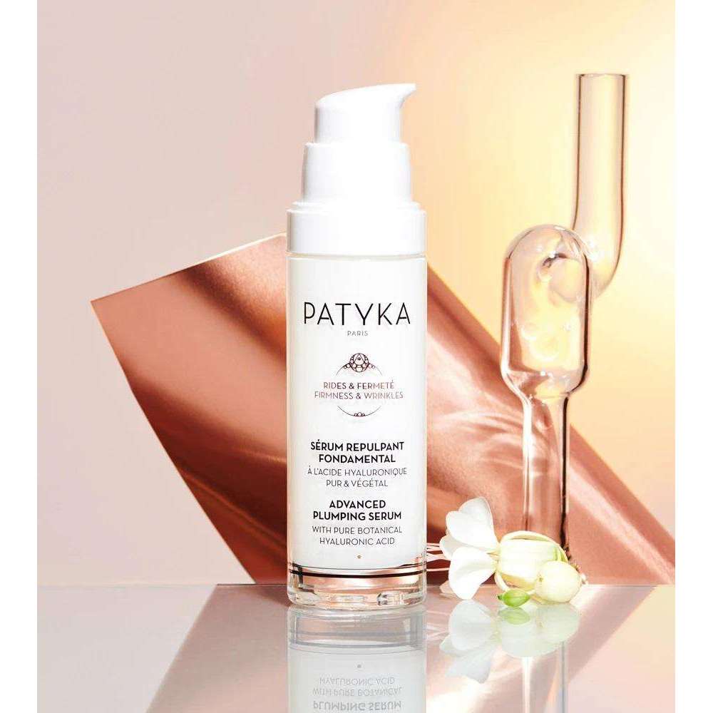 Patyka Advanced Plumping Serum -pullo pystyssä mattapersikanvärisellä alustalla; pullo sylinterimäinen, kirkasta tai huurrettua lasia, valkoinen korkki; etiketissä "Patyka Advanced Plumping Serum" yksinkertaisella mustalla fontilla; lähellä kirkkaita lasikoriste-esineitä ja pieniä valkoisia kukkia; tausta jaettu persikan- ja beigensävyihin; ei ihmisiä näkyvissä.
