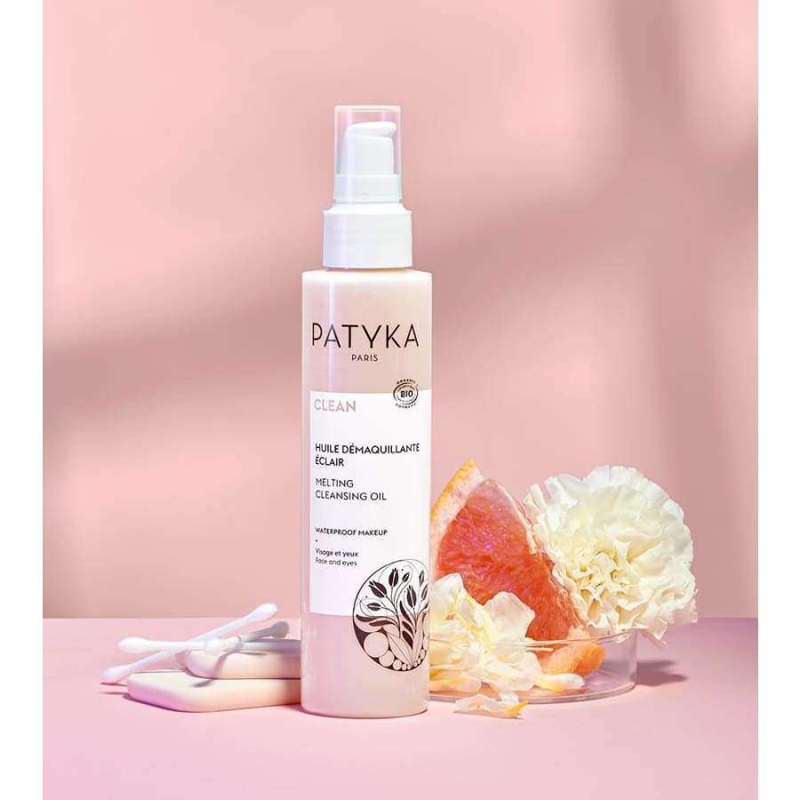 Patyka Melting Cleansing Oil -pullo, kirkas sylinterimäinen pullo valkoisella pumppukorkilla, pystyssä mattapinkillä alustalla; pullossa vähäistä mustaa tekstiä ja ohutta tekstausvapaata etikettiä; ympärillä useita valkoisia pumpulipuikkoja, halkaistuja vaaleanpunaisia greippiviipaleita, yksi valkoinen kukka näkyvin terälehdin ja irrallisia kukanterälehtiä; tausta vaaleanpunainen läpi kuvan.