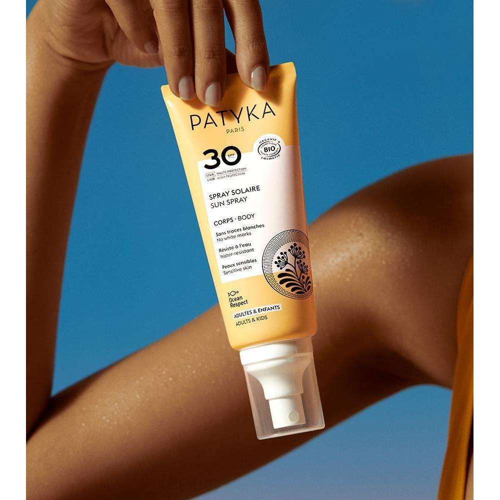 Patyka Sun Body Milk SPF30; käsi pitelee keltaista, mattapintaista tuubia tekstillä "PATYKA Paris SPF 30 Sun Spray"; pakkaus lieriömäinen ja muovinen yksinkertaisella groteskitypografialla; etiketti kertoo laajakirjoisesta suojasta vartalolle sekä sopivuudesta aikuisille ja lapsille; osa käsivarrresta näkyvissä; tausta yhtenäisen sininen.