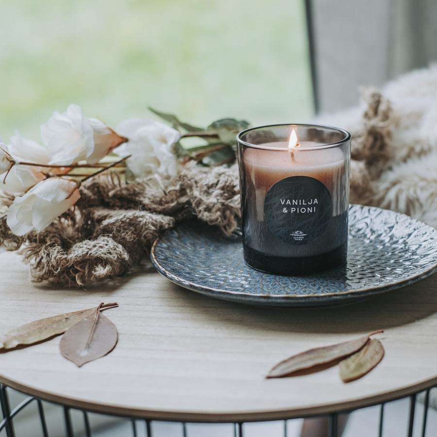 Saaren Taika Decor TuoksukynttilÀ Vanilja & Pioni; sytytetty kynttilÀ tekstillÀ "VANILJA & PIONI" tekstuurisella sinisellÀ keraamisella lautasella, vierellÀ kuivattuja kukkia ja eukalyptuksenlehtiÀ; oikealla osittain nÀkyvissÀ taitettu neulottu huopa; puinen alusta, taustalla ikkunasta tulevaa pÀivÀnvaloa; etiketin teksti yksinkertaisella mustalla groteskikirjasimella valkoisella pohjalla; ei ihmisiÀ tai kÀsiÀ kuvassa.