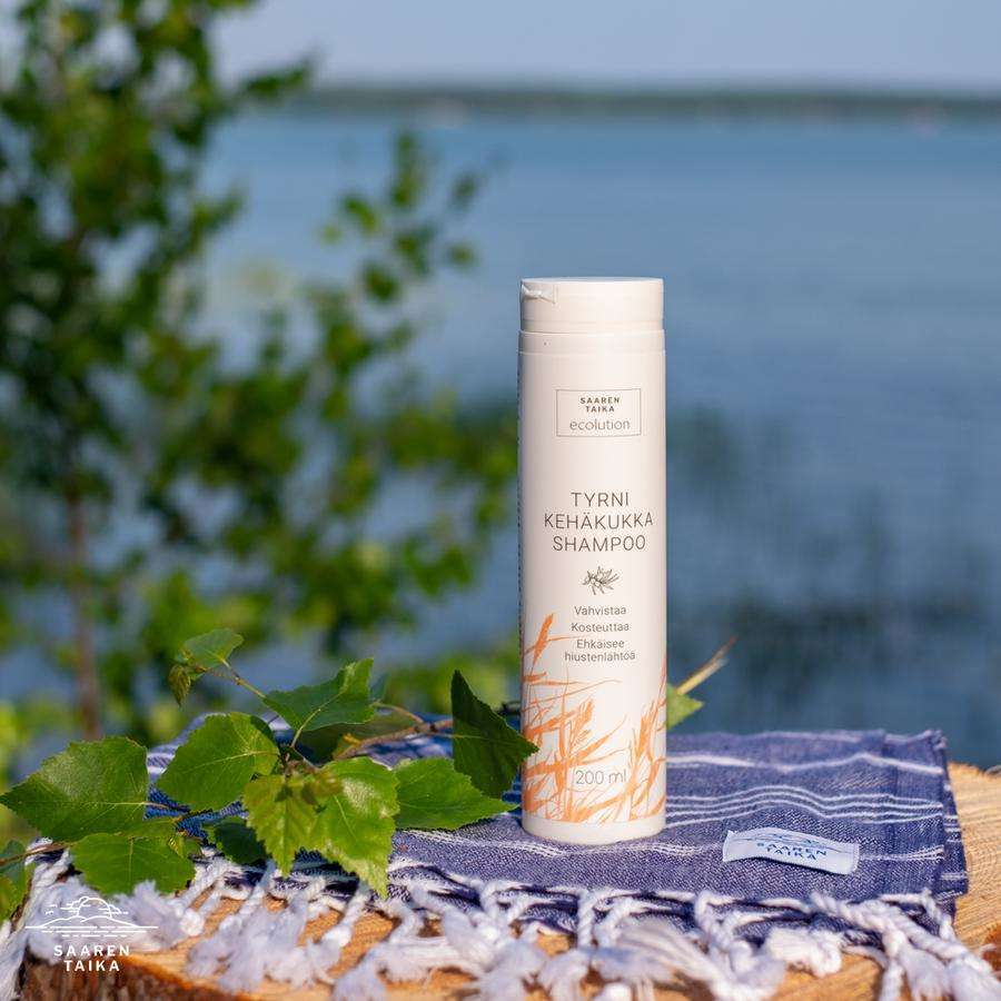 Saaren Taika Ecolution Shampoo Tyrni-Kehäkukka; pullo pystyssä raidallisella pyyhkeellä ulkona järven rannalla; pullo sylinterinmuotoinen, opaakki tai kevyesti sävytetty, etiketissä luettavissa teksti "Tyrni Kehäkukka"; vihreitä lehtiä etualalla, taustalla auringonvaloa heijastavaa tyyntä vettä ja sininen taivas; ei ihmisiä tai käsiä kuvassa.