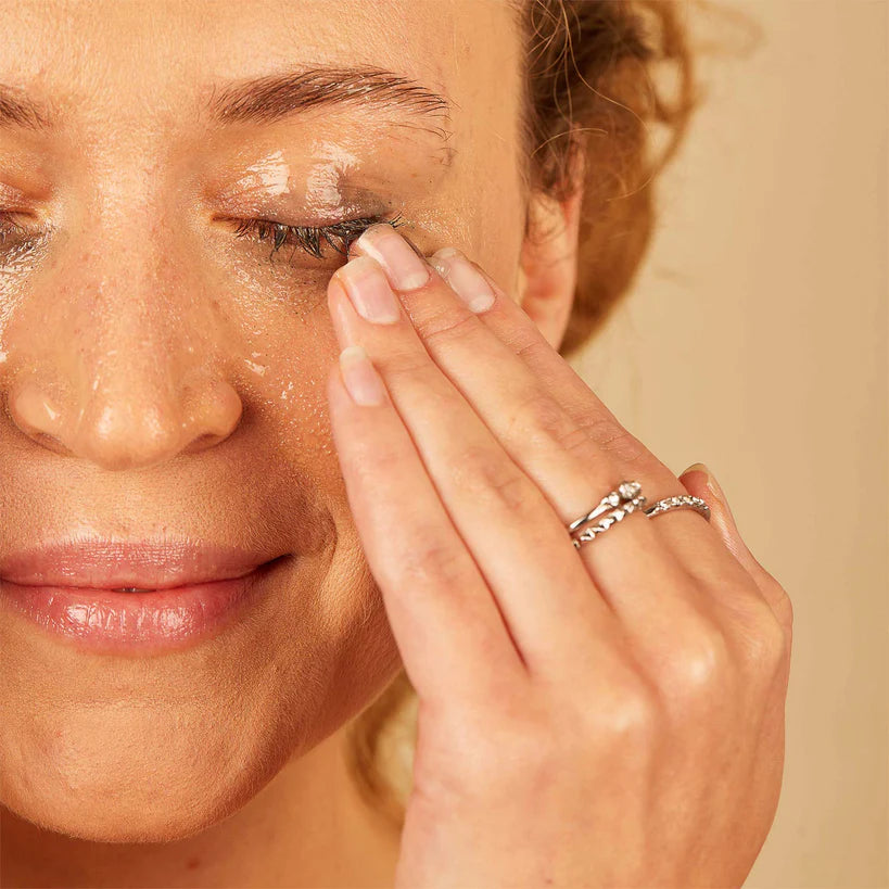 Skin & Tonic Calm Clean puhdistusbalmi; henkilö hierolee puhdistusainetta suljetulle silmäluomelleen sormenpäillään, silmät kiinni ja lievä hymy, iho kimmeltää kosteutettuna; ei näkyvää pakkausta; lähikuva kasvoista ja käsistä neutraalilla taustalla.