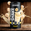 3K Protein Shake Banana; sylinterin muotoinen Layenberger 3K Protein Shake -purkki banaanin makuisena vaalealla puupinnalla; pakkaus keltainen ja valkoinen kiiltopinnalla, suljettu muovikannella; etiketissä maininnat ”glutenfrei” ja ”24 g proteiinia”; purkin ympärillä maitoroiskeita ja useita banaaniviipaleita; ei ihmisiä tai käsiä kuvassa.
