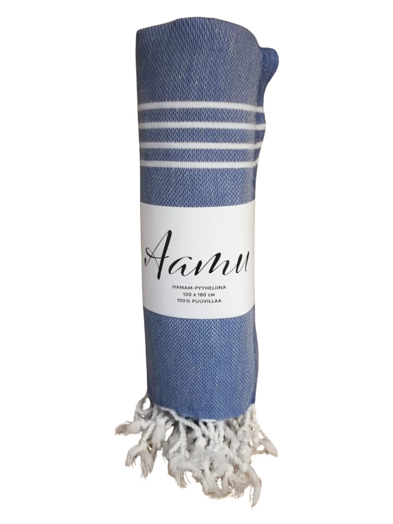 AAMU Hamam Kylpypyyhe; kääritty sini-valkoinen pyyhe valkoisin hapsuin alaosassa, kääritty etiketillä, jossa lukee "Aanu"; pyyhkeessä vuorottelevia sinisiä ja valkoisia raitoja, materiaali kudottua kangasta mattapinnalla; etiketti paperia yksinkertaisella sans-serif-tekstillä; tausta yksinkertainen ja siisti; ei ihmisiä tai käsiä kuvassa.