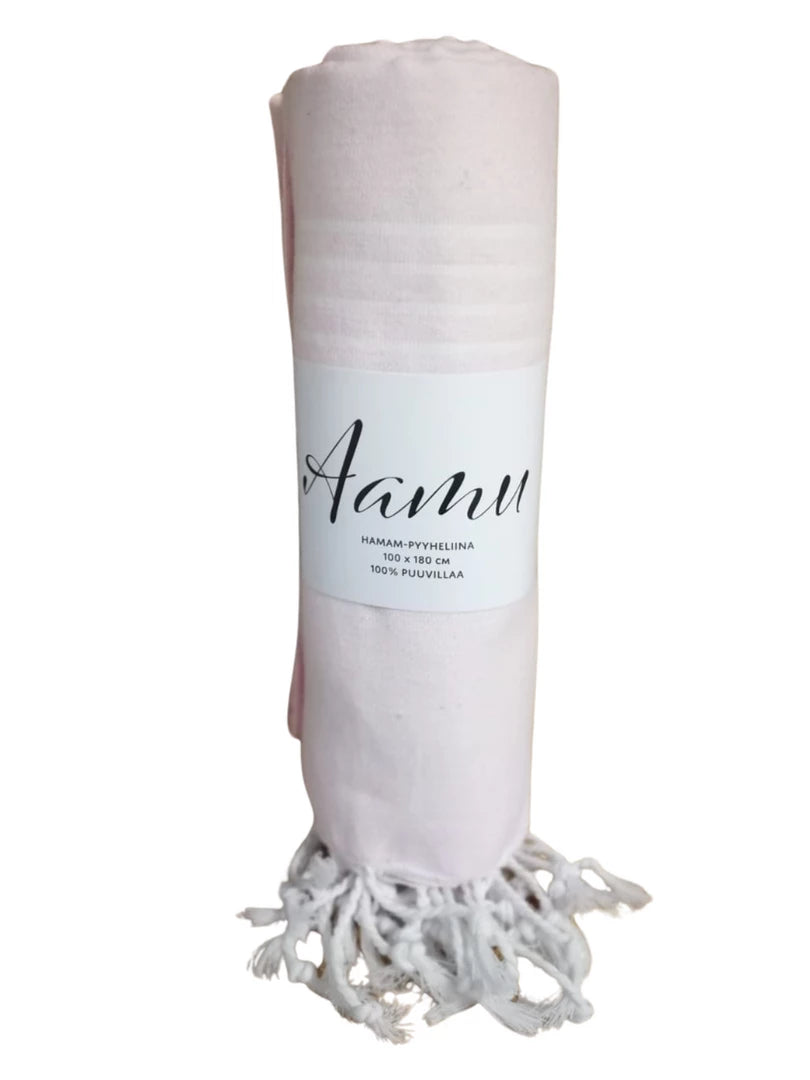 AAMU Hamam Kylpypyyhe; kääritty vaaleanpunainen puuvillapyyhe valkoisin hapsuin päissä; pyyhe kääritty etiketillä, jossa lukee "Aanu" ja "100% cotton" suomeksi selkeällä sans-serif-fontilla; materiaalin pinta mattainen ja tekstiilimäinen; ei ihmisiä tai käsiä näkyvissä; tausta yksinkertainen ja siisti; keskikuva lähietäisyydeltä.