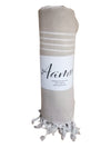 Beige rolled-up Aamu towel with white stripes and fringed ends, labeled on a white wrapper. The wrapper displays the “Aamu” name in a simple sans-serif font. The towel is cylindrical in shape, matte textured fabric, no visible branding on the towel itself. Photo taken against a plain white background, no people or props present.