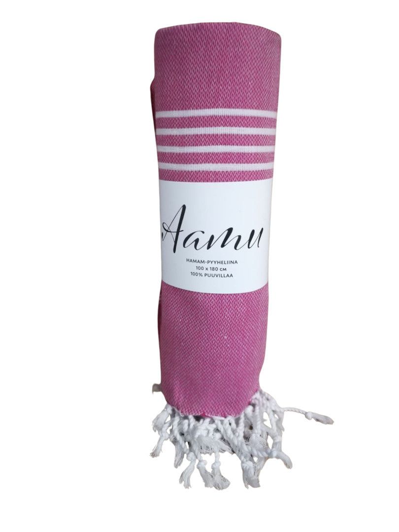 A rolled-up pink towel with white stripes and white fringe, wrapped in a label that reads Aamu and additional text. The towel is cylindrical, features a matte fabric finish, and has straight edges with visible tassel detailing at each end. The product sits on a plain light background. No people or hands are present in the image.