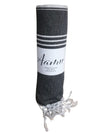 A dark gray rolled towel with a matte finish, featuring white fringe along one edge and two parallel white stripes near the top. The towel is made of cotton and is labeled “Aamu” in simple sans-serif font; label also states “100% cotton.” Product is unboxed, photographed on a neutral surface. No people or hands present.
