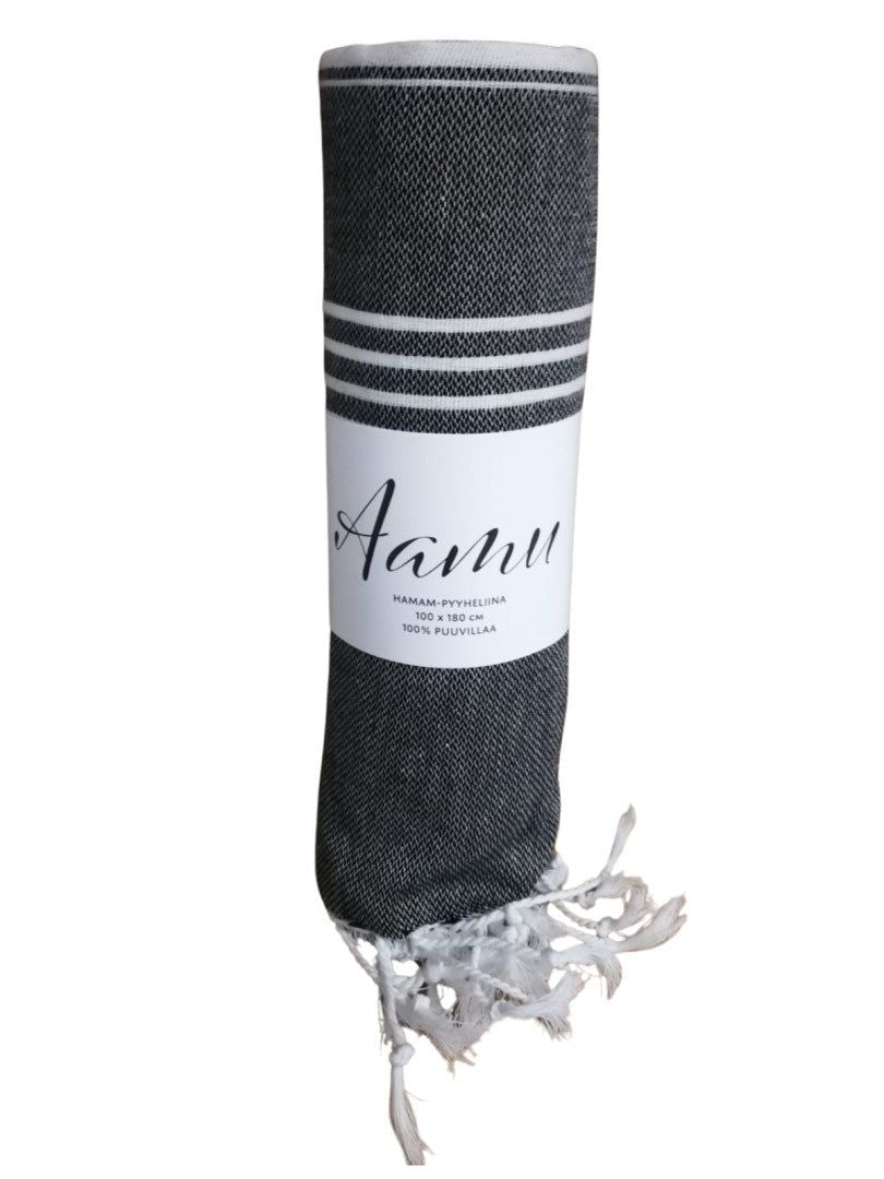 A dark gray rolled towel with a matte finish, featuring white fringe along one edge and two parallel white stripes near the top. The towel is made of cotton and is labeled “Aamu” in simple sans-serif font; label also states “100% cotton.” Product is unboxed, photographed on a neutral surface. No people or hands present.