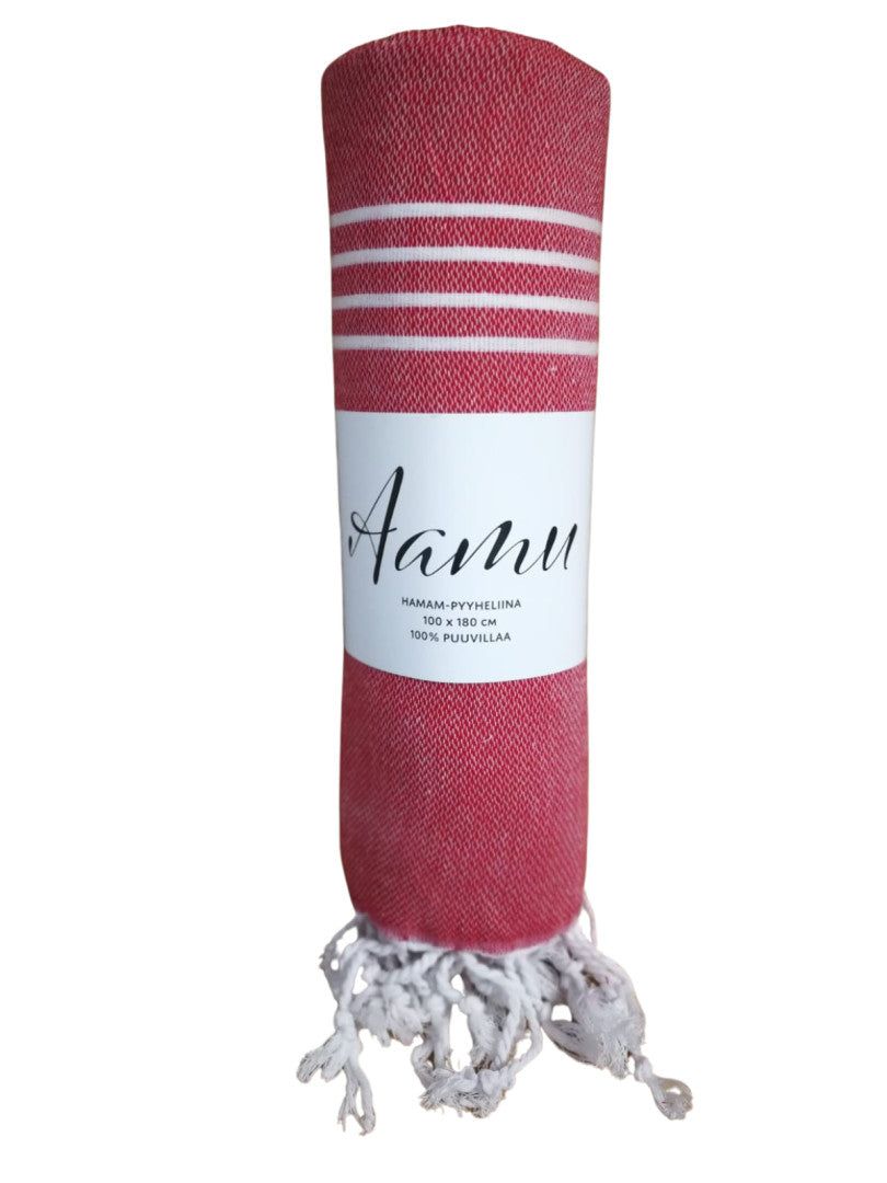A rolled red towel with distinct white stripes and a small rectangular white fabric label showing the text “Aanu” in simple black lettering. The towel is textile, matte-surfaced, cylindrical when rolled, with white twisted fringe along one end. It stands upright on a solid plain white background. No people or packaging are visible in the photo.