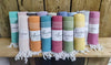 Ten rolled, colorful, striped towels with white fringe are displayed upright on a wooden shelf. Each towel features a white label printed with the word “Aamu” in simple sans-serif type. The scene is set against a weathered wooden wall. No packaging other than the labels is visible; no people or hands are present in the image.