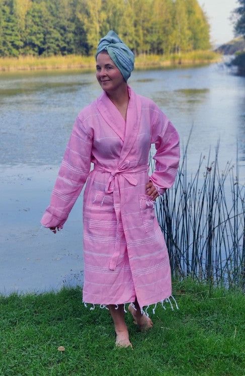 AAMU Hamam Kylpytakki; henkilö vaaleanpunaisessa kylpytakissa ja vihreä pyyhe päässä seisoo paljain jaloin ruoholla rauhallisen joen äärellä, ympärillä ruokoja ja puita; kohtaus on ulkona luonnonvalossa; kuvassa ei näy pakkausta tai brändäystä; vain ruohoa näkyy pintana, ei typografiaa tai etikettitekstiä; koko vartalon kuva maan tasalta.