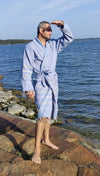 A man in a blue striped robe stands barefoot on the rocky shore by the water, shading his eyes with one hand and gazing out over the shimmering surface on a sunny day. The robe is long, loose-fitting, with horizontal stripes in varying shades of blue and white. The fabric appears lightweight and matte, with no visible logos or text. Outdoor setting shows rocks, water, sunlight reflections. No packaging present; no other people or props visible. Full-body shot from side angle.