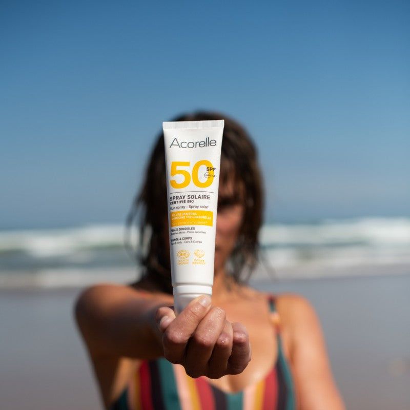 Acorelle Aurinkosuojasuihke Kasvoille ja Vartalolle SPF50: rannalla oleva henkilö ojentaa Acorelle SPF 50+ -aurinkovoidetuubia, taustalla meri ja taivas pehmeästi epätarkkoina; valkoinen muovituubi on mattapintainen ja pyöristetyin reunoin; etiketissä oranssi-harmaa groteskiteksti "Acorelle" ja "SPF 50+"; henkilön käsi näkyy selvästi etualalla.