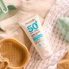 Acorelle Baby Aurinkovoide SPF 50+: tuubi vaaleanvihreillä rannan leluilla koristellulla tekstuuripintaisella kankaalla; tuubi on valkoinen ja mattapintainen, oranssi etiketti groteskikirjasimella tekstillä "baby" ja "SPF 50+"; vieressä pastellivihreitä muovisia rantaleluja, yksi simpukan ja toinen lehden muotoinen; ei ihmisiä tai käsiä kuvassa; pehmeä luonnonvalo.