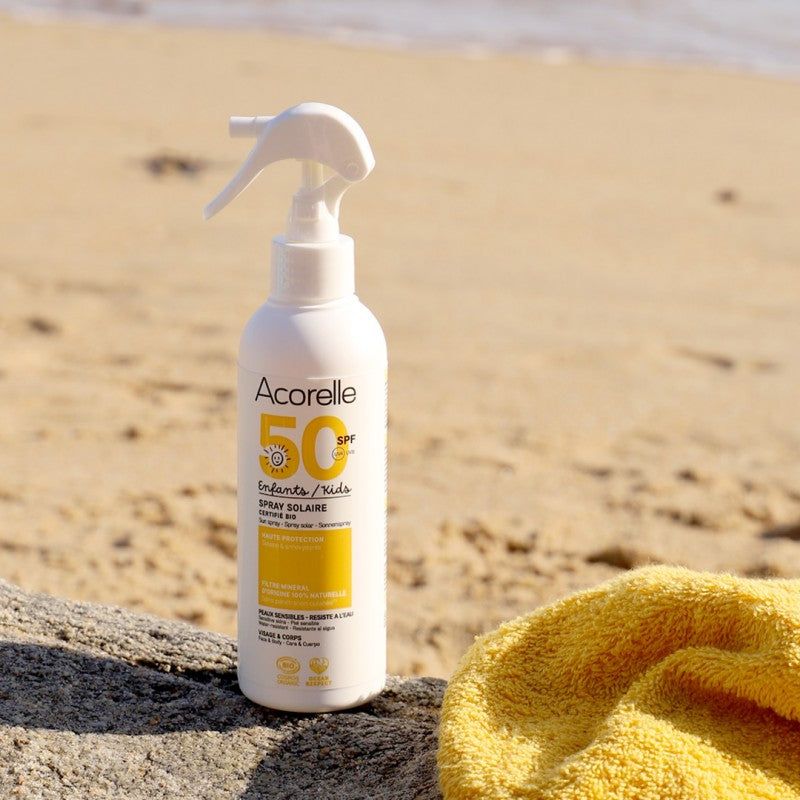 Pullo Acorelle SPF 50 -luomuaurinkosuojasuihketta seisoo pystyssä hienolla hiekalla, vieressä siististi taiteltu keltainen pyyhe. Muovipullo on valkoinen oranssilla korkilla, lieriömäinen ja mattapintainen. Etiketissä lukee brändinimi Acorelle yksinkertaisella pääteviivattomalla fontilla ja SPF 50 lihavoituna. Taustalla näkyy sumeana meri ja sininen taivas.