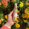 Acorelle Vartalotuoksu Fresh & Fruity: käsi pitelee sylinterimäistä läpinäkyvää suihkepulloa valkoisella nokalla ja läpinäkyvällä korkilla; etiketissä teksti "Acorelle Eau d'Été" modernilla groteskikirjasimella sekä pienempää tekstiä alla; pullo ja nokka ovat kiiltäväpintaisia; taustalla kukkivia keltaisia ja oransseja kukkia ulkona luonnonvalossa; lähikuva, käsi näkyvissä.
