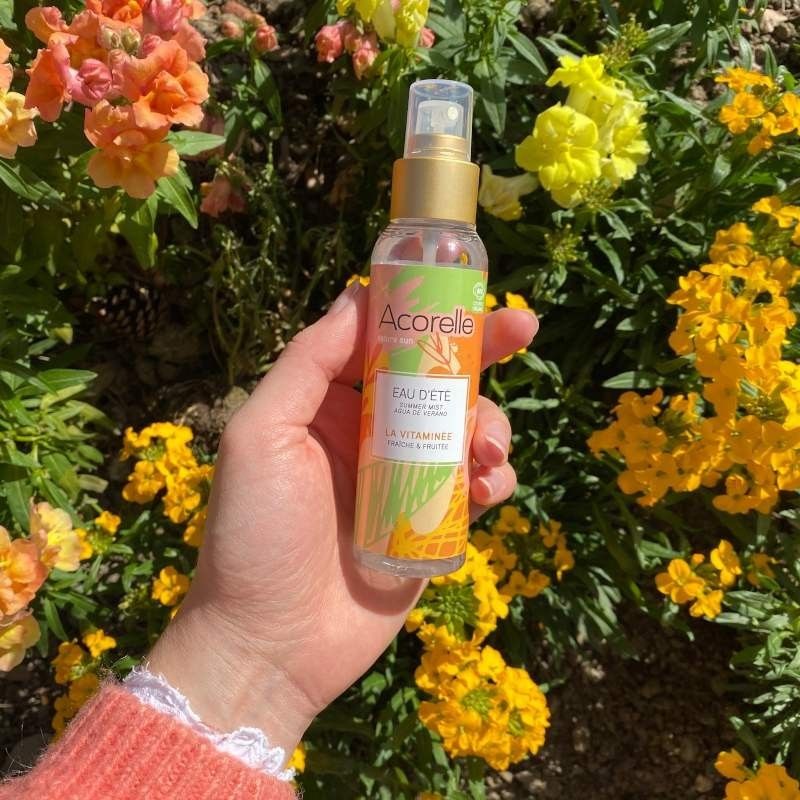 Acorelle Vartalotuoksu Fresh & Fruity: käsi pitelee sylinterimäistä läpinäkyvää suihkepulloa valkoisella nokalla ja läpinäkyvällä korkilla; etiketissä teksti "Acorelle Eau d'Été" modernilla groteskikirjasimella sekä pienempää tekstiä alla; pullo ja nokka ovat kiiltäväpintaisia; taustalla kukkivia keltaisia ja oransseja kukkia ulkona luonnonvalossa; lähikuva, käsi näkyvissä.