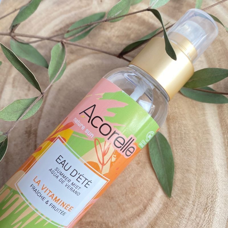 Acorelle Vartalotuoksu Fresh & Fruity: Eau d'Été -kesäsuihkepullo suorakulmaisella läpinäkyvällä rungolla ja sylinterimäisellä korkilla, monivärinen kiiltävä etiketti tekstillä "Acorelle" ja "Eau d'Été" selkeällä modernilla kirjasimella; pullo seisoo sileällä puupinnalla suurten tummanvihreiden lehtien ympäröimänä; ei ihmisiä tai käsiä kuvassa.