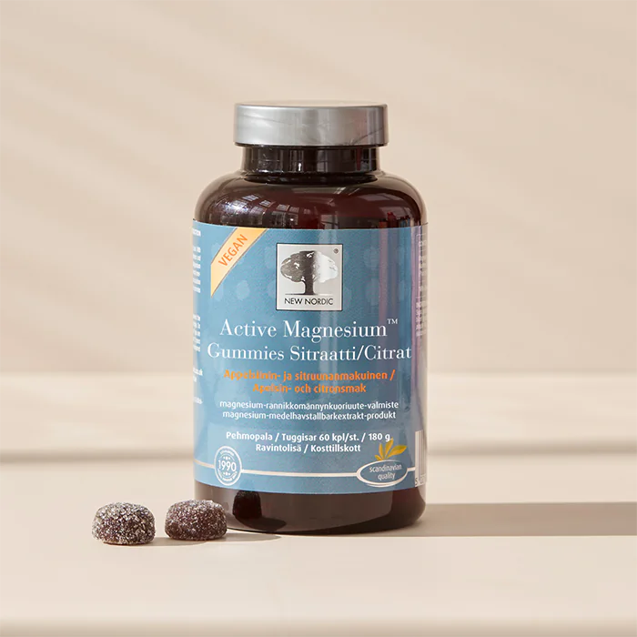 Active Magnesium Gummies Sitraatti; pullo New Nordic Active Magnesium Citrate Vegan Gummies seisoo beigellä pinnalla, kaksi magnesiumpurutablettia vieressä; pullo lieriömäinen muovia valkoisella kannella; etiketti sininen, näyttää luettavaa valkoista ja oranssia tekstiä groteskifontilla; ei ihmisiä tai käsiä; asetelma yksinkertainen ilman muita esineitä kuvassa.