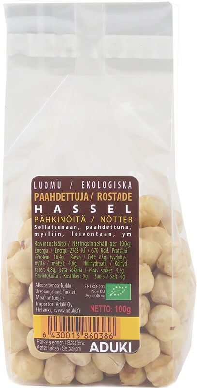 Aduki Luomu Paahdettu Hasselpähkinä; kirkas muovipussi Aduki-merkkisiä luomu-paahdettuja hasselpähkinöitä, suorakaiteen muotoinen, mattapintainen etiketti näyttää suomen- ja ruotsinkielistä tekstiä, ravintosisältötaulukon, viivakoodin ja nettopainon 100 g; etiketissä groteskifontti mustana valkoisella pohjalla; ei ihmisiä tai käsiä; neutraali, tavallinen tausta lähikuvassa.
