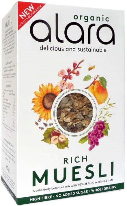 Alara Organic Rich Muesli -laatikon etusivu, jossa eloisia hedelmä- ja siemenkuvituksia, kirkas ikkuna näyttää tuotteen sisällön sekä luettavia merkintöjä: runsaskuituinen, täysjyvää, ei lisättyä sokeria; laatikko suorakaiteen muotoinen mattapinnalla; groteskikirjasinta käytetty läpi pakkauksen; tausta yksinkertainen ja siisti; ei ihmisiä tai käsiä kuvassa.