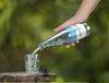 A hand pours sparkling water from a clear, cylindrical glass bottle labeled ALKUWAT into a transparent glass containing several ice cubes. The bottle has a glossy finish. The label features simple, bold text. A loose bottle cap lies next to the glass on a hard surface. The scene is set outdoors with an out-of-focus green background in natural sunlight.