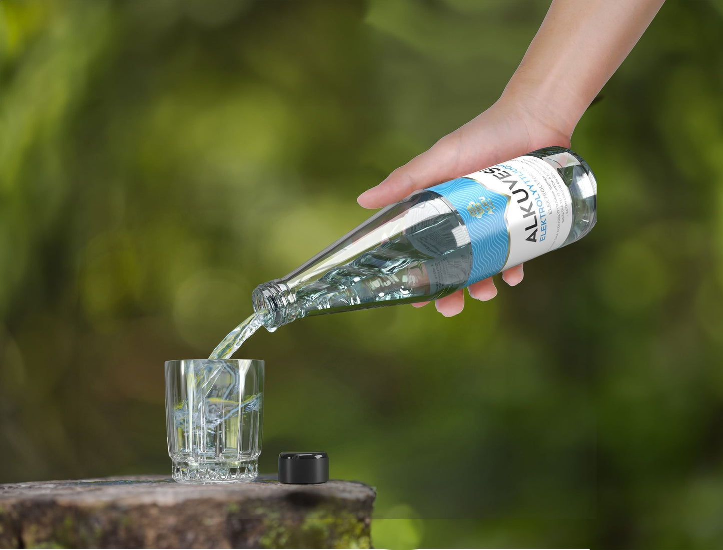 A hand pours sparkling water from a clear, cylindrical glass bottle labeled ALKUWAT into a transparent glass containing several ice cubes. The bottle has a glossy finish. The label features simple, bold text. A loose bottle cap lies next to the glass on a hard surface. The scene is set outdoors with an out-of-focus green background in natural sunlight.
