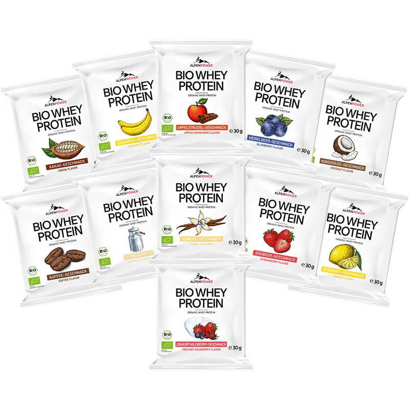 AlpenPower Luomu Heraproteiini Vanilja; kymmenen Bio Whey Protein -annospussia eri makuina, mukaan lukien suklaa, banaani, metsämarjat, kookos, cappuccino, vanilja, mansikka ja mango, aseteltuna riveihin valkoiselle taustalle; suorakulmaiset pussit mattapinnalla painetuin etiketein, joissa brändi- ja makunimet lihavoidulla groteskikirjasimella; ei ihmisiä tai käsiä; kuva otettu ylhäältäpäin.