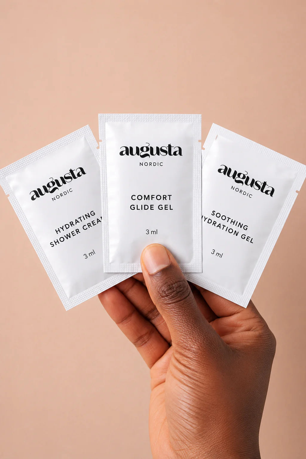 Käsi pitelee kolmea Augusta Nordic -näytepussia — Hydrating Shower Cream, Comfort Glide Gel ja Soothing Hydration Gel, kukin 3 ml — neutraalia beigeä taustaa vasten. Pussit ovat suorakulmaisia, mattapintaisia, ja niissä on selkeästi painetut tuotenimet pääteviivattomalla fontilla. Kuvassa näkyvät vain käsi ja näytepussit; muita esineitä ei näy.