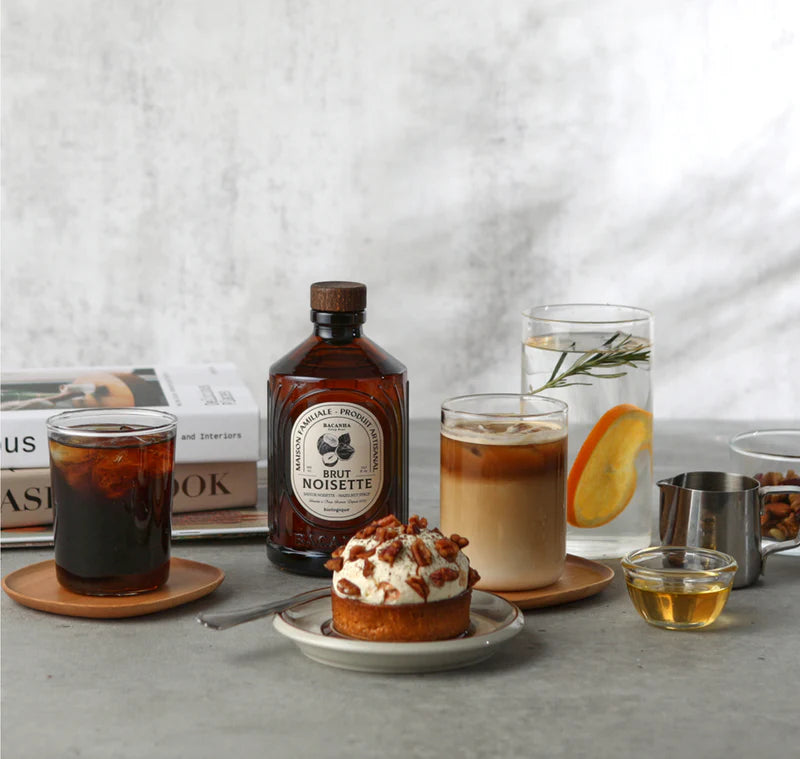 Bacanha Luomu Hasselpähkinäsiirappi; pöytä, jolla lasisia ja posliinisia kahvijuomia, kirkas pullo tekstillä Brut Noisette mustalla serif-fontilla, pähkinöillä koristeltu pyöreä jälkiruoka, appelsiiniviipalein ja rosmariinioksin täytetty vesitumma, kuusikulmainen hunajapurkki ilman brändiä, mantelein täytetty valkoinen kuppi ja pinottuja kirjoja taustalla luovat viihtyisän tunnelman.