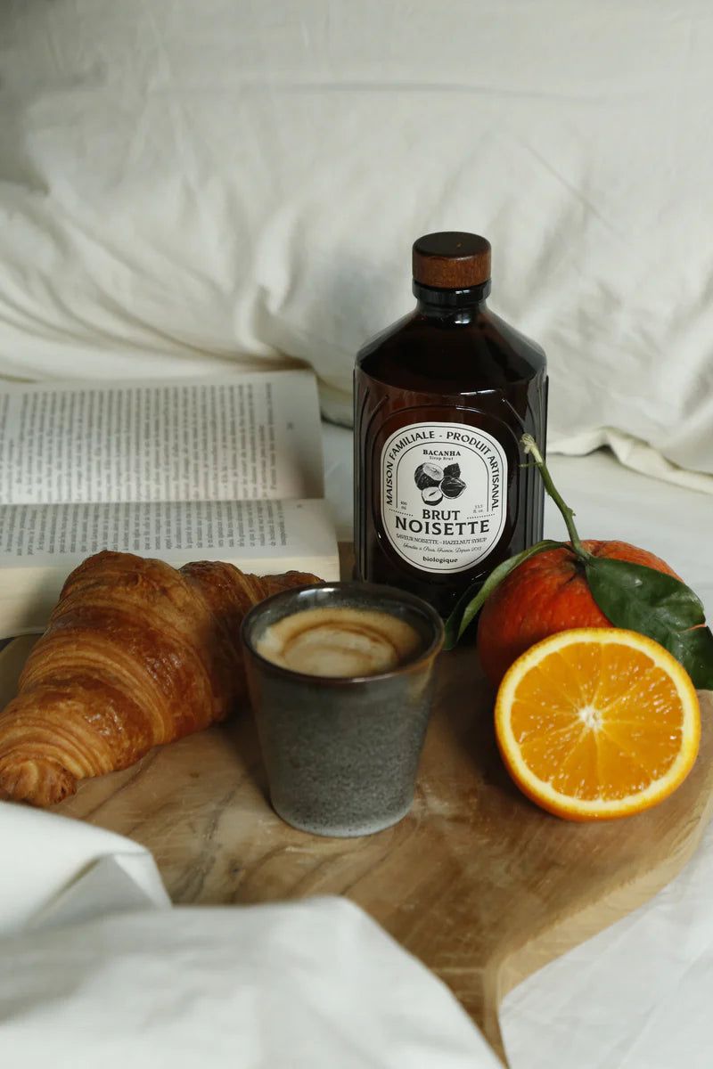 Bacanha Luomu Hasselpähkinäsiirappi; croissant, kahvimuki, puolikas ja kokonainen appelsiini sekä kirkas pullo tekstillä Brut Noisette mustalla serif-fontilla, aseteltuna suorakaiteen muotoiselle puutarjottimelle; tarjotin avoimen kirjan vieressä ryppyisillä valkoisilla lakanoilla; ei ihmisiä tai käsiä; kuvattu yläviistosta.