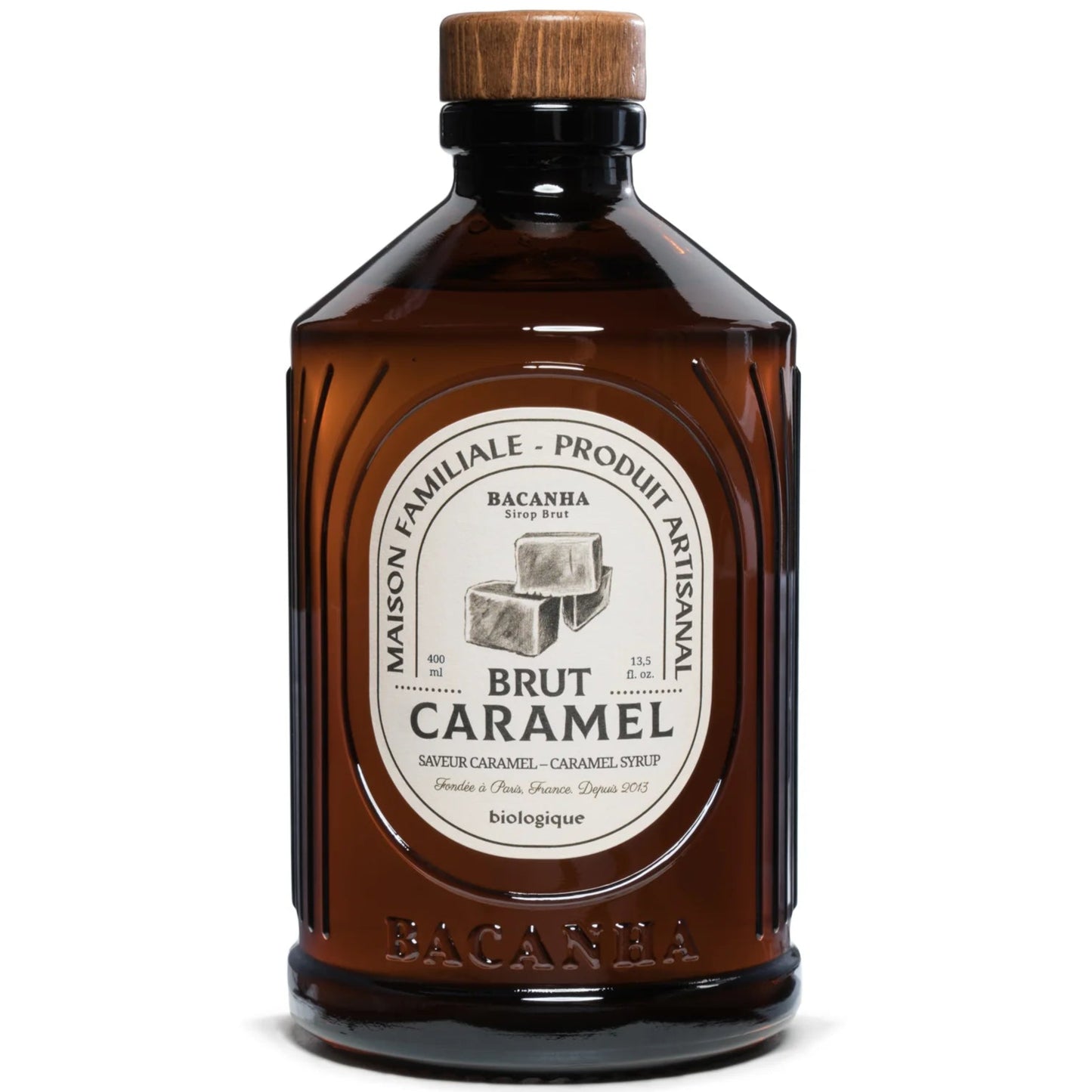 Bacanha Luomu Karamellisiirappi; ruskea lieriömäinen lasipullo litteällä puukorkilla; etiketissä teksti Brut Caramel syrup by Bacanha sekä karamellipalojen kuvia; maininnat luomu, käsityöläistuote ja täyteläinen karamellin maku; lihavoitu musta ja kultainen groteskifontti; kuvattu valkoisella taustalla ilman ihmisiä tai käsiä.